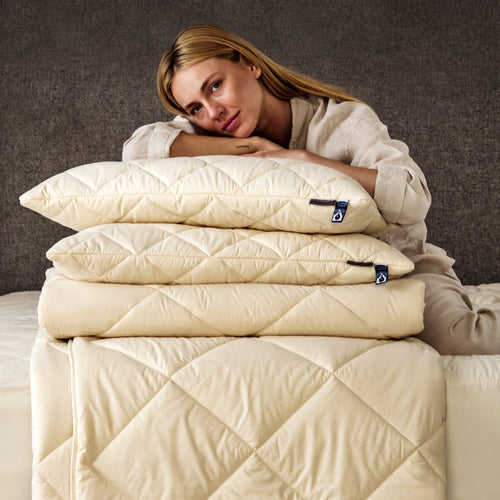 A blonde lady sitting on a mattress, with a pile of Woolroom bedding, including a duvet, pillows and mattress protector