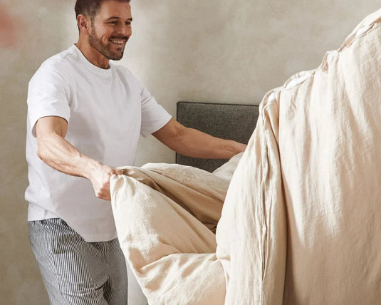 Man in a white t-shirt and gray pants making a bed with beige bedding.