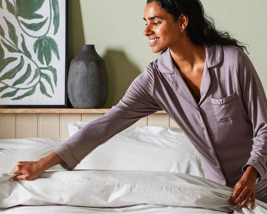 Woman in a purple shirt standing by a bed with white bedding.