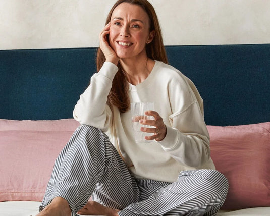 Woman sitting on a bed holding a glass, wearing a white sweatshirt and striped pants.